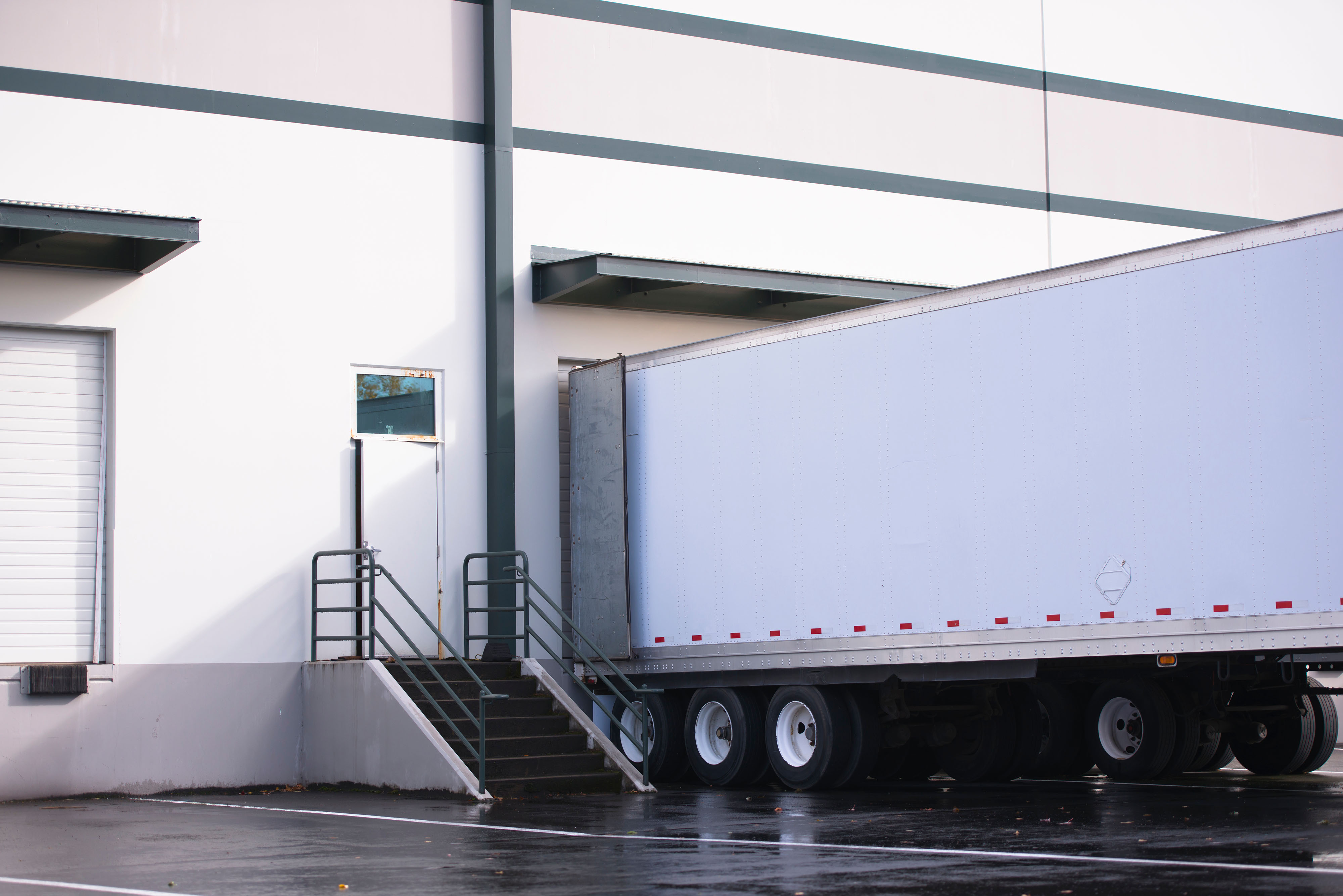 Warehouse loading dock, box truck backed up to bay door, workers unloading freight, industrial overhead lighting