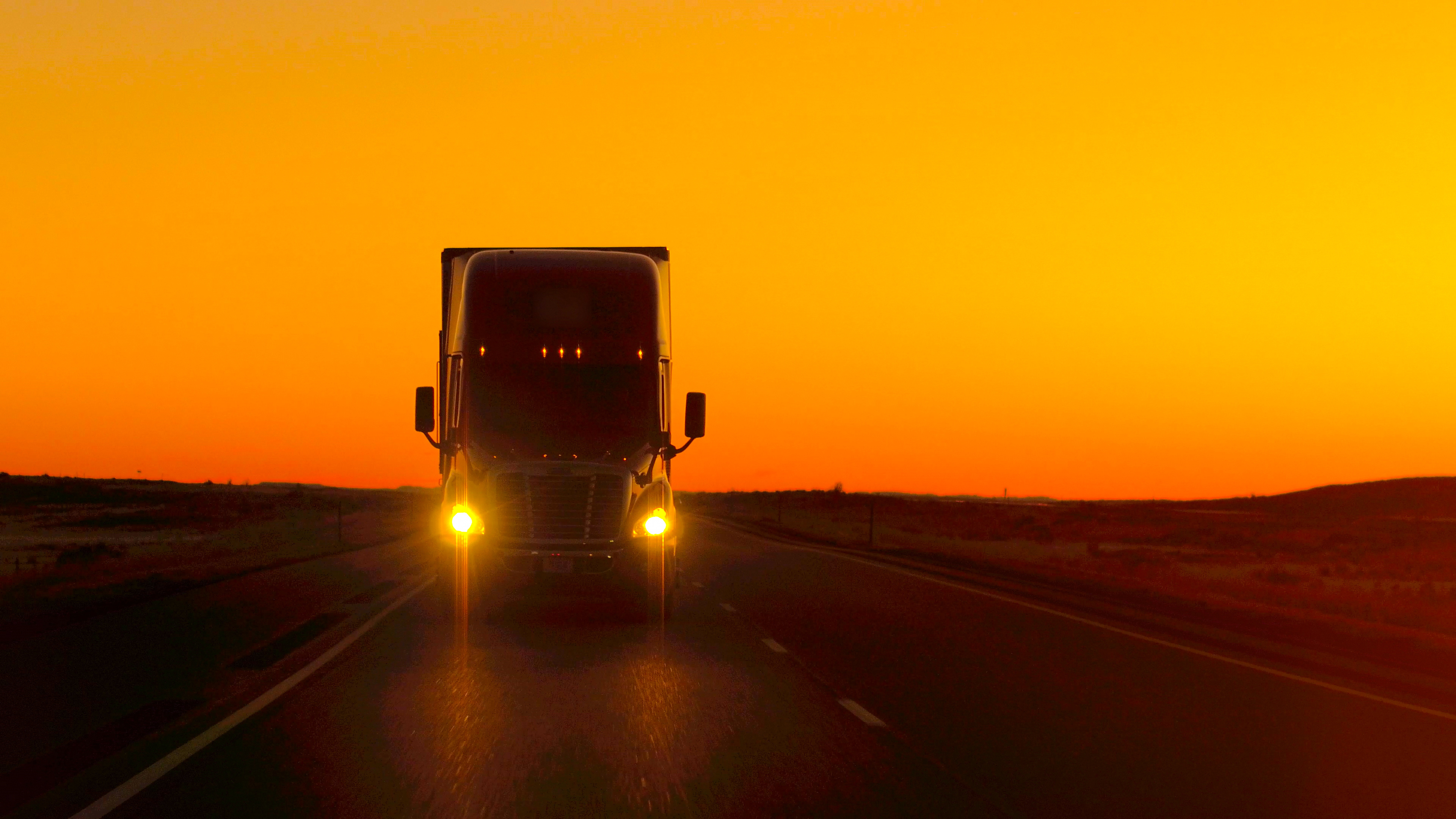 Cargo truck on a highway at dusk, lights on, wide open road ahead, cinematic dark tone